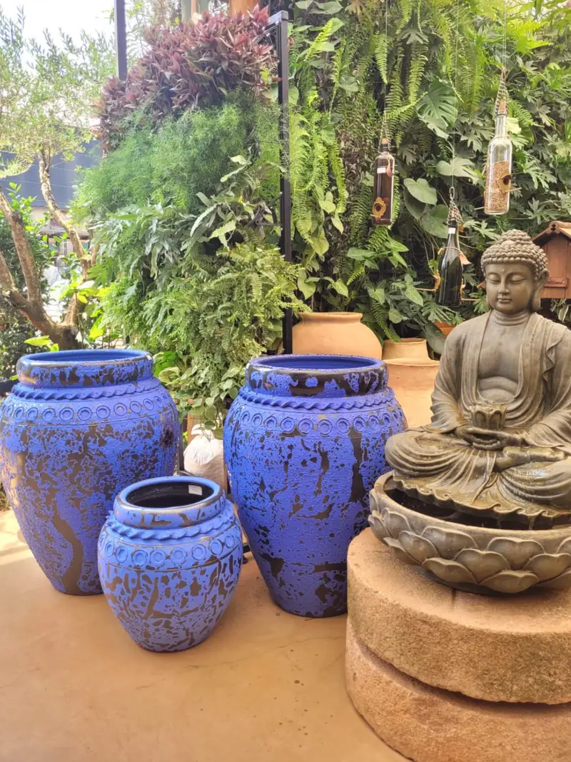 Vasos vietnamitas azuis e escultura de Buda em pedra, expostos sob a vegetação do pátio da loja