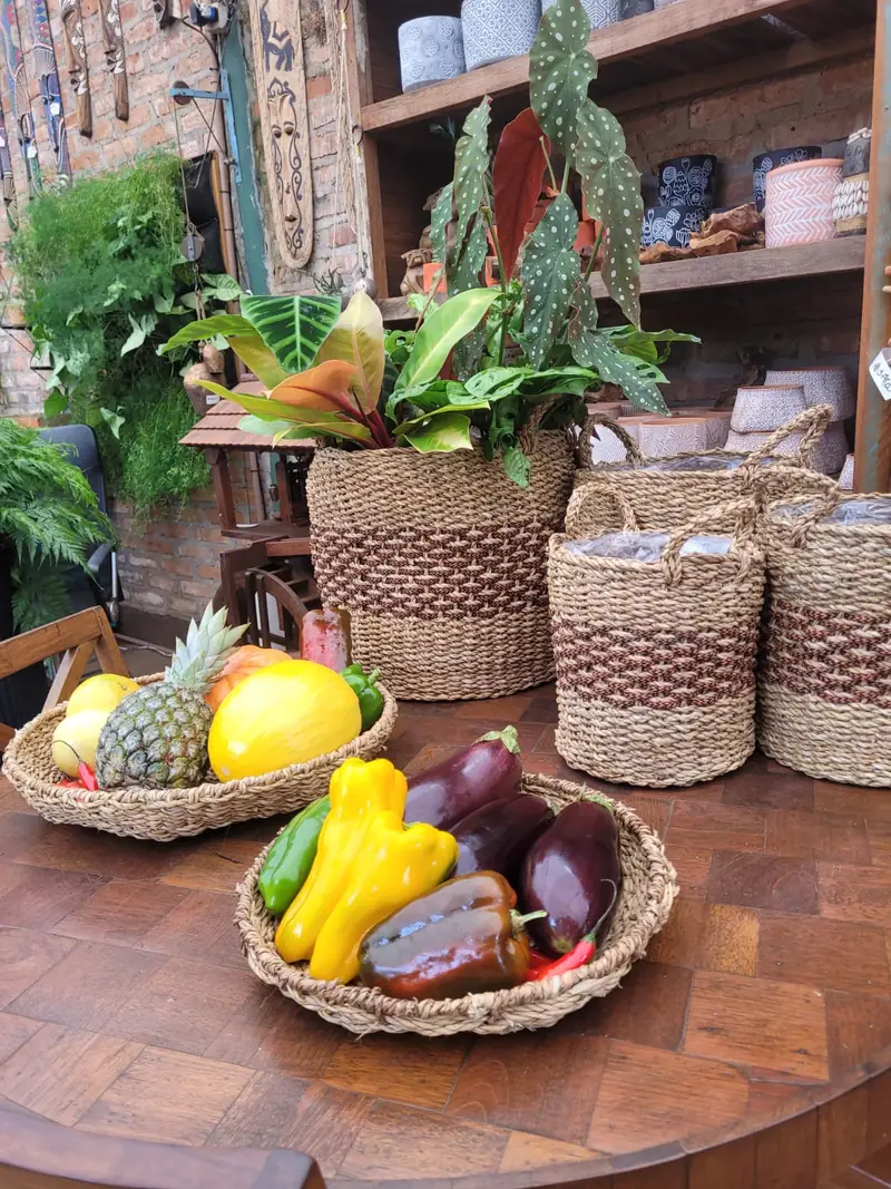 Arranjo de folhagens em cesto de palha sobre mesa de madeira, com cestos de fibra natural e um bowl de frutas ao lado