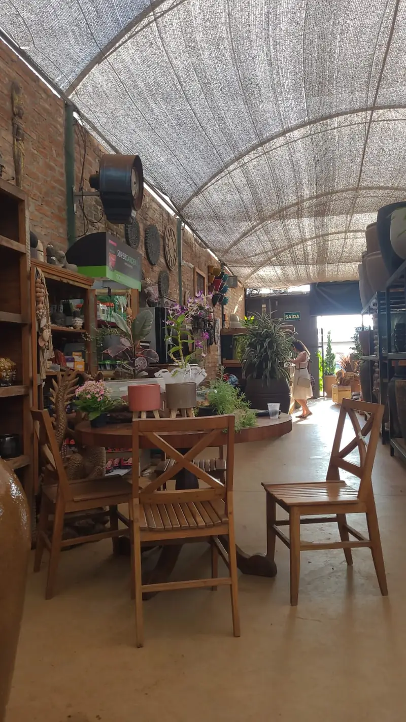 Corredor coberto da loja, com mesa e cadeiras de madeira e plantas dos dois lados