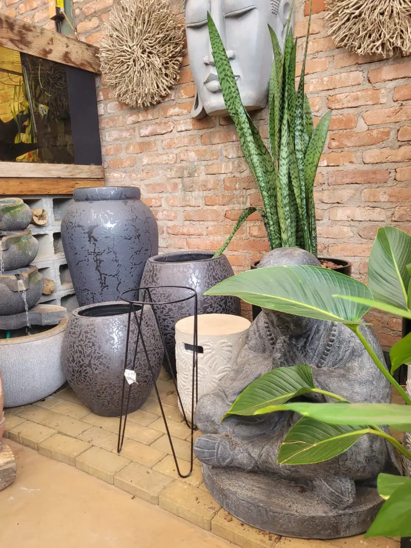 Vasos altos de cimento e cerâmica escura expostos junto a uma parede de tijolinho, com sansevieria e bananeira-ornamental
